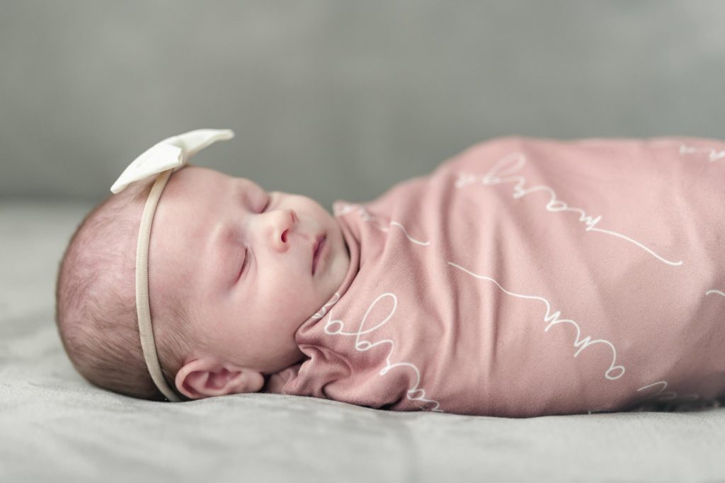 A newborn baby wears a white bow and is wrapped in a pink swaddle with her name on it.