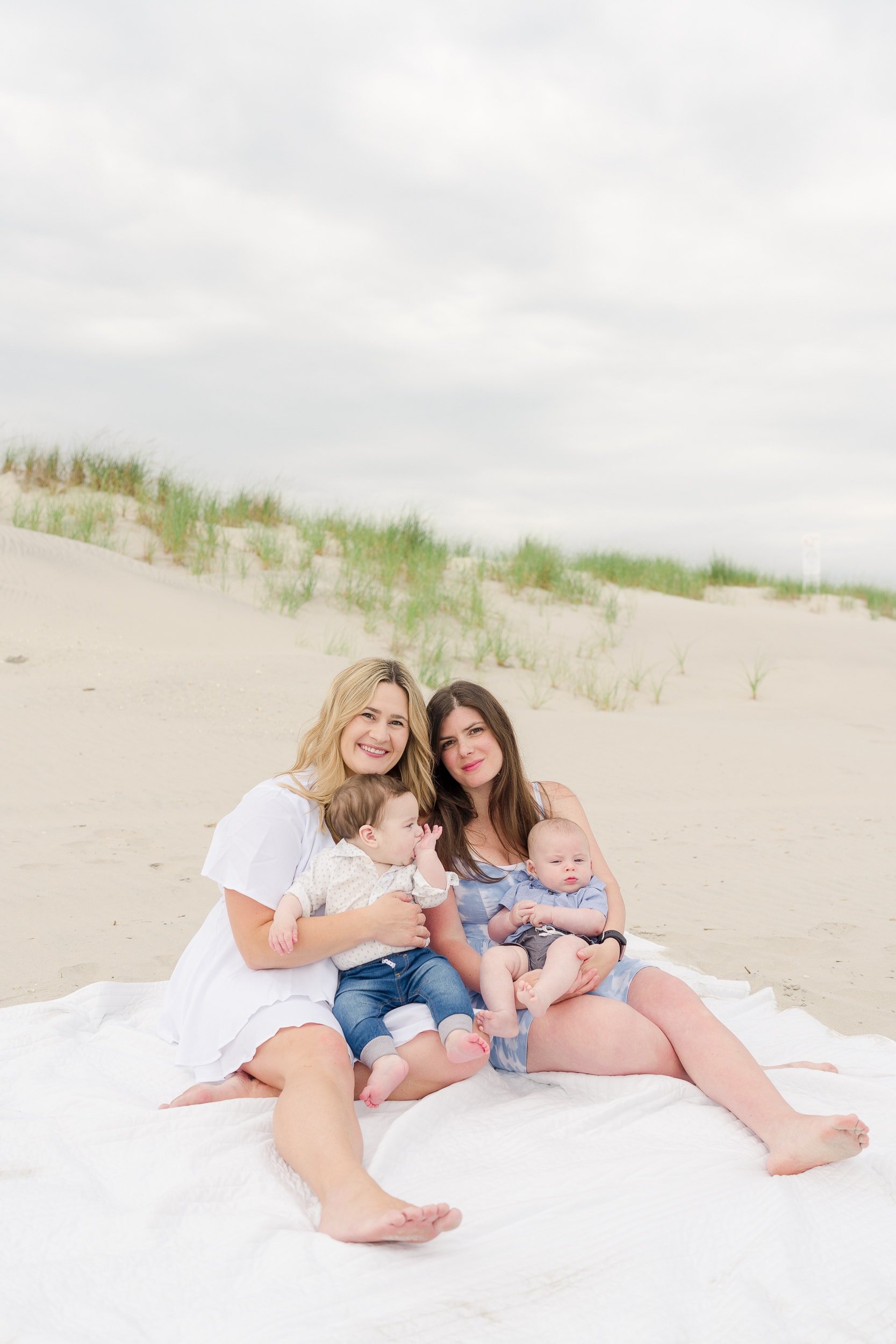 Two sisters sit on a white blanket at the beach in Avalon with their newborn babies.
