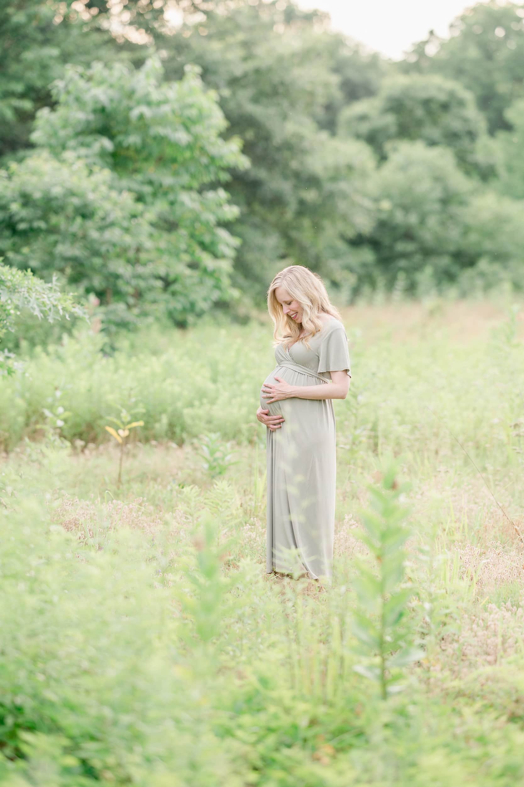 A pregnant woman wearing a green dress poses in a field during her summer maternity photoshoot.