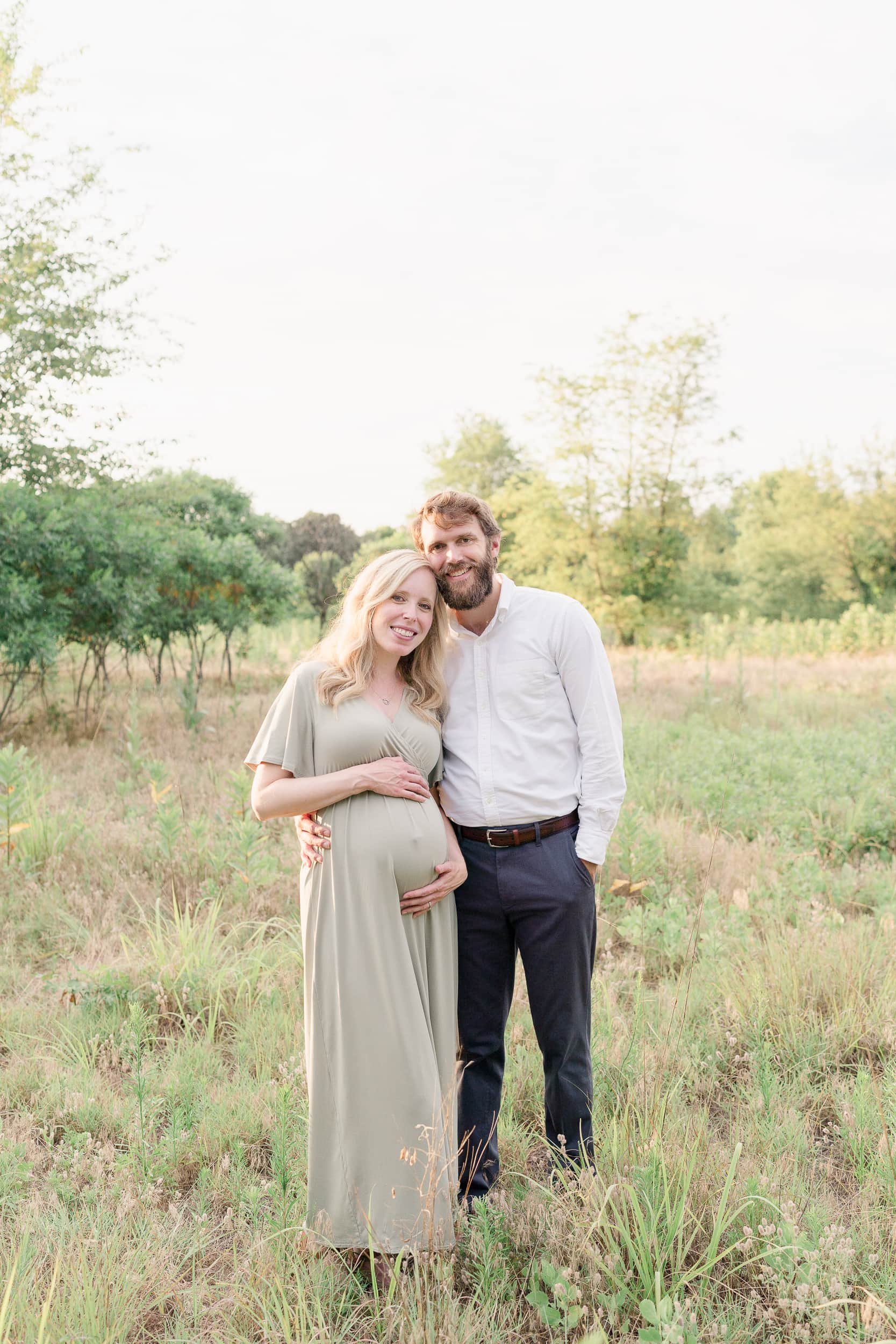 A smiling portrait of a pregnant woman and her husband standing in a field by Moorestown Maternity Photographer Courtney Landrum.