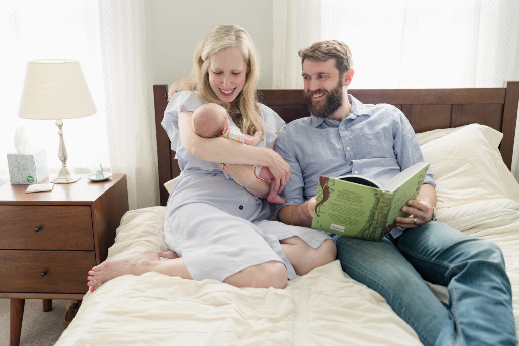 New parents read a book to their newborn daughter while sitting in bed.