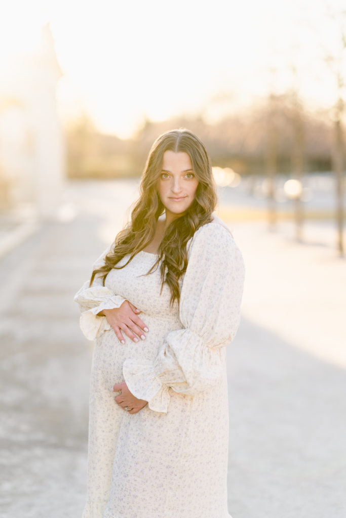 Pregnant woman holds her belly and stares into the camera while the winter sun shines around her.
