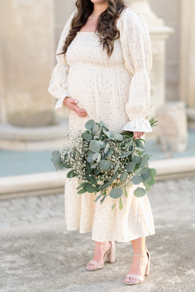 Pregnant woman holds her belly and a bouquet of eucalyptus and baby's breath.