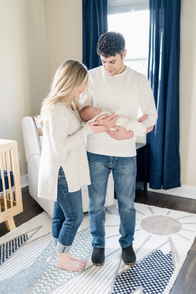 Mother and father wearing white sweaters stand in the nursery room while holding their baby boy.