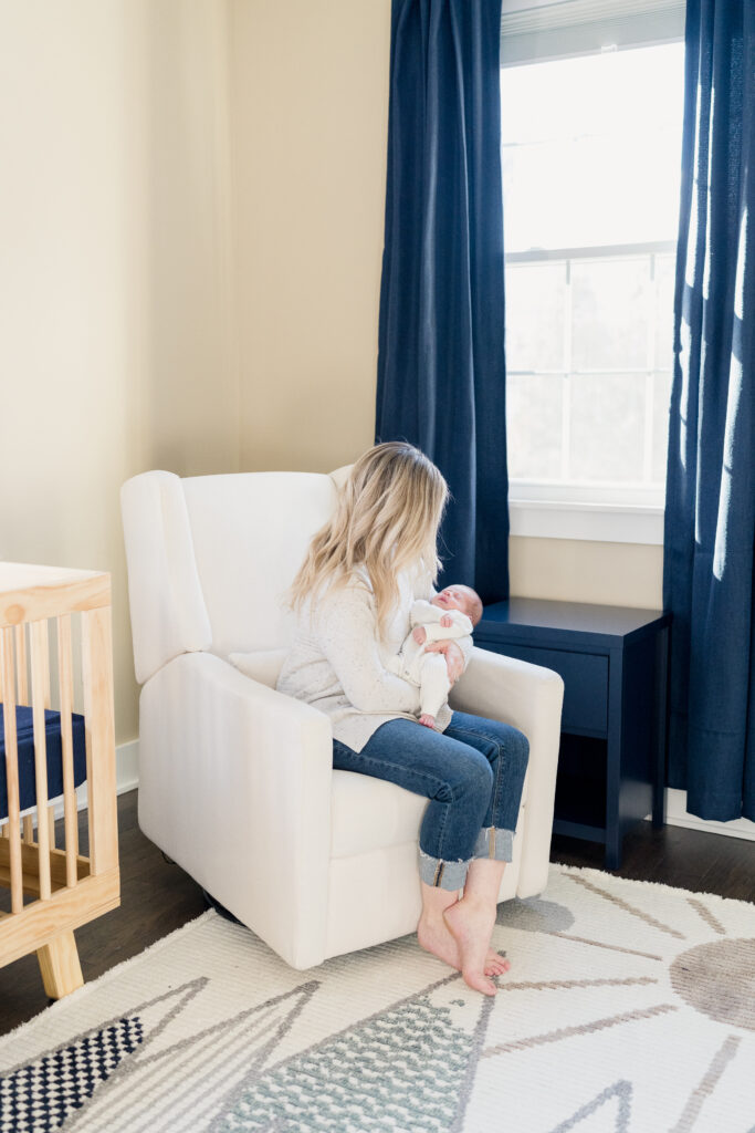 Mom sits in a white chair in a navy and white nursery with her newborn son.