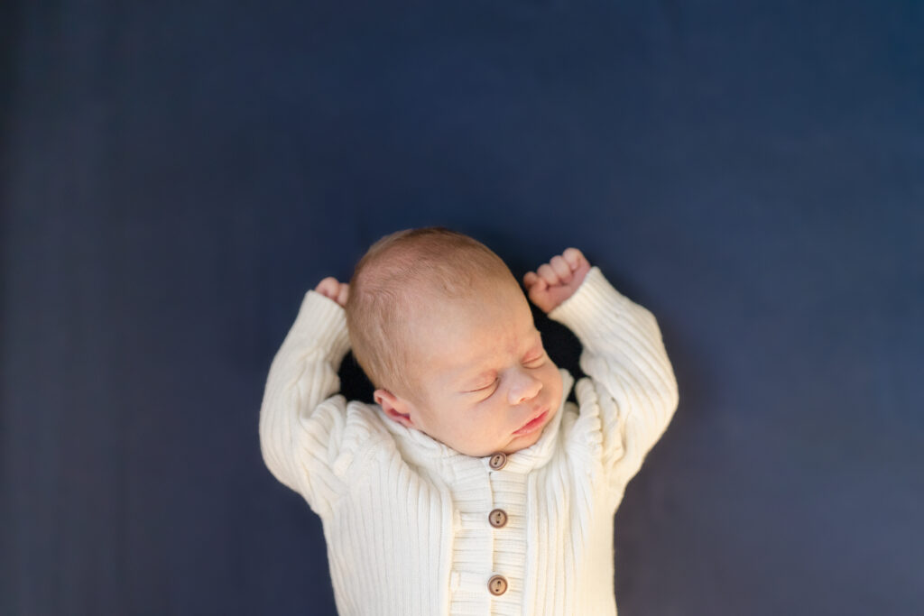 A newborn baby boy wearing a white sweater laying on a navy blanket.