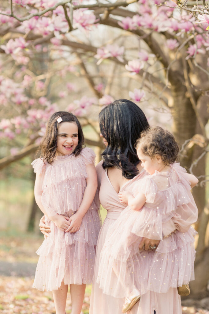 A mom and her daughters wearing pink dresses stand under a magnolia tree in full bloom, smiling at each other.