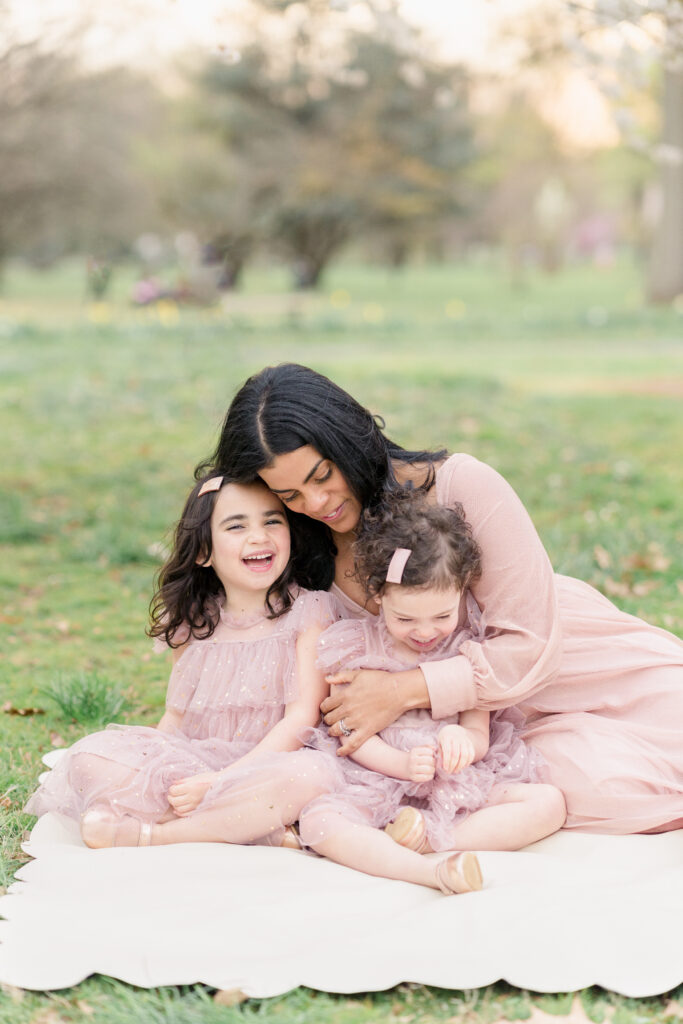Mother and two daughters sitting on a white gathre blanket wearing pink dresses.