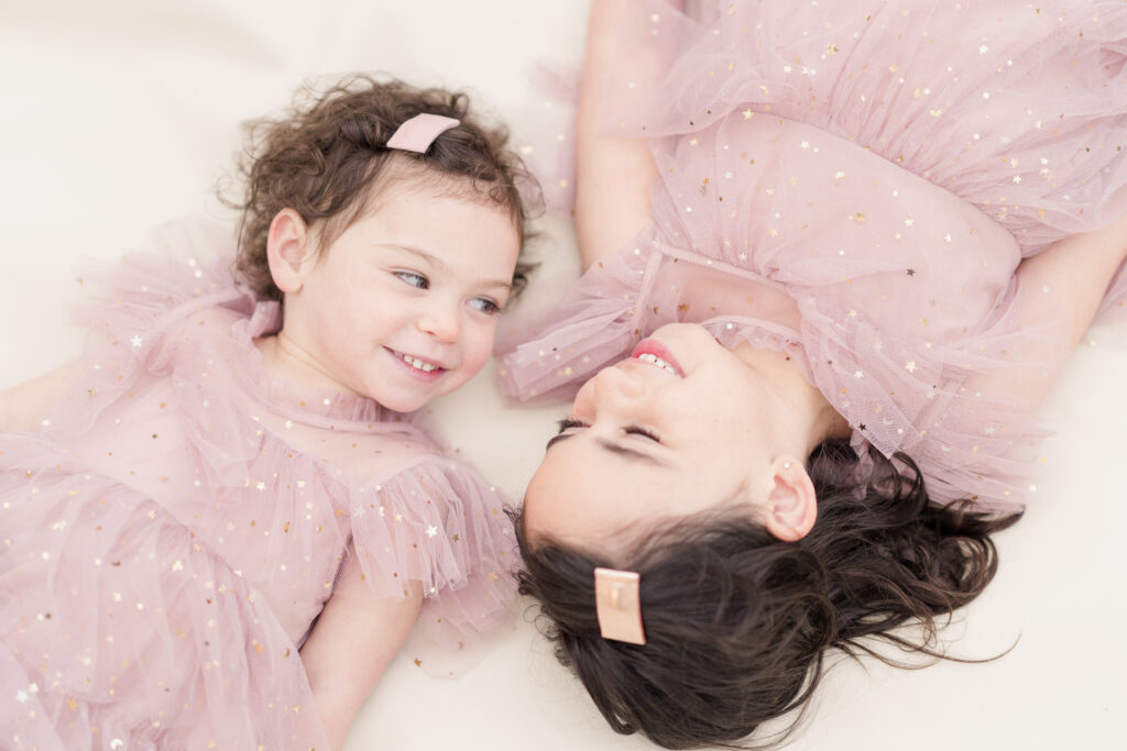 Two sisters lay on a white blanket, looking at each other and smiling.