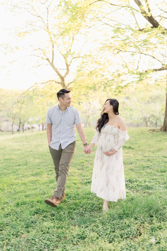A pregnant couple holds hands and walks across a field, looking at each other and laughing.
