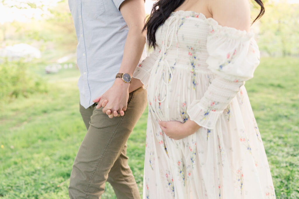 A close-up of a pregnant woman holds her belly while holding her husband's hand.