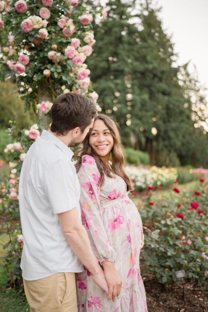 A woman 5 months pregnant stands with her husband in front of a lamp post covered in roses.