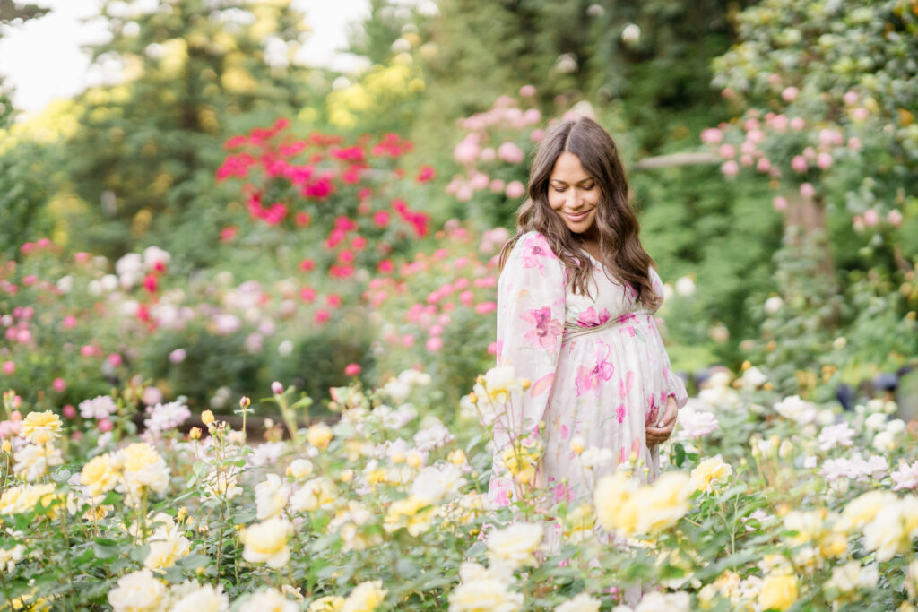 A pregnant woman stands in a garden of roses in the summer.