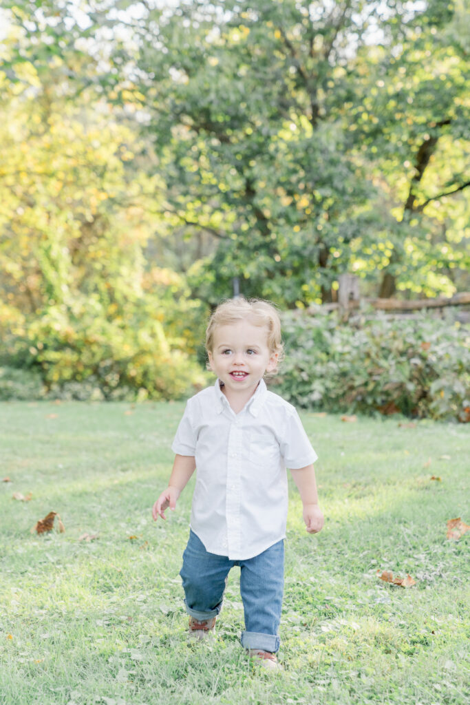 Little boy in a white button-up and jeans walks toward the camera and smiles.
