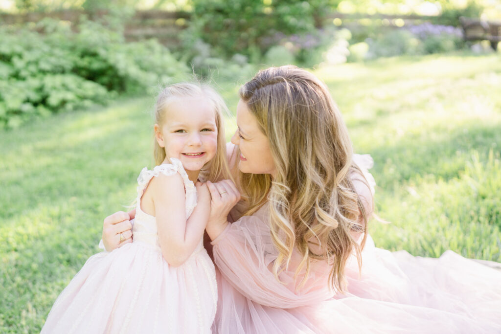 Little girl in a pink dress smiles at the camera with her mom.