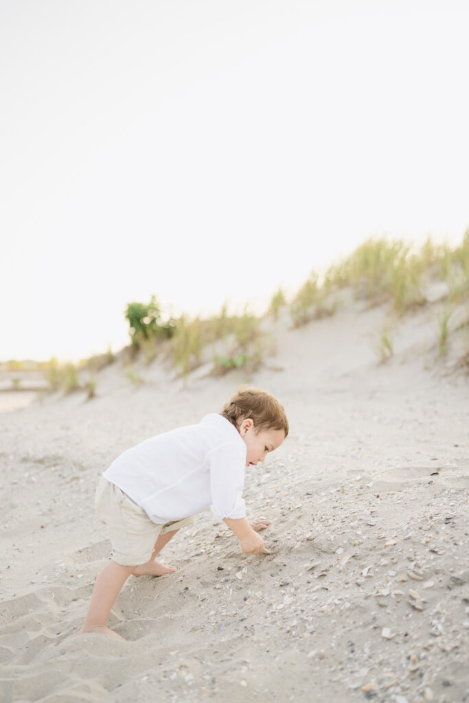 A baby boy wearing a white shirt and khaki shorts digs in the sand for seashells at the Jersey shore. 