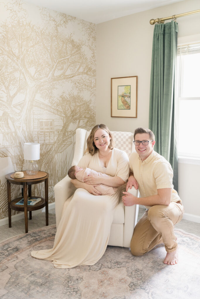 Mother and father sit in their beige and sage nursery and smile while holding their newborn girl in their arms.