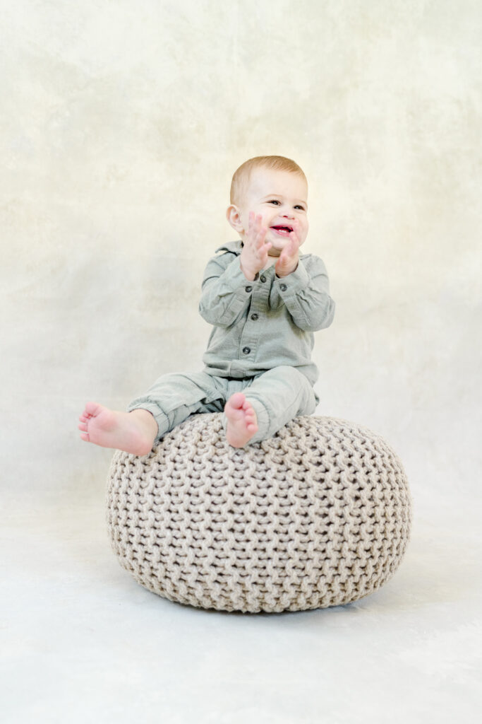 A baby in a green jumpsuit claps his hands while sitting on a gray poof.