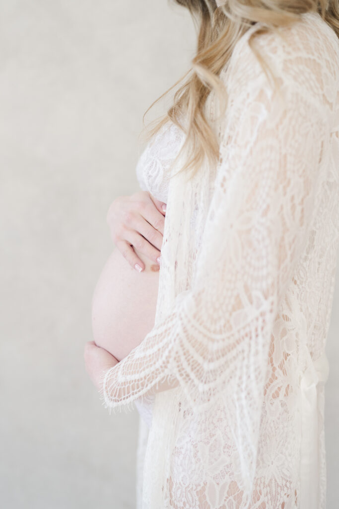 A mother holds her belly while wearing a white lace robe in a studio maternity photoshoot.