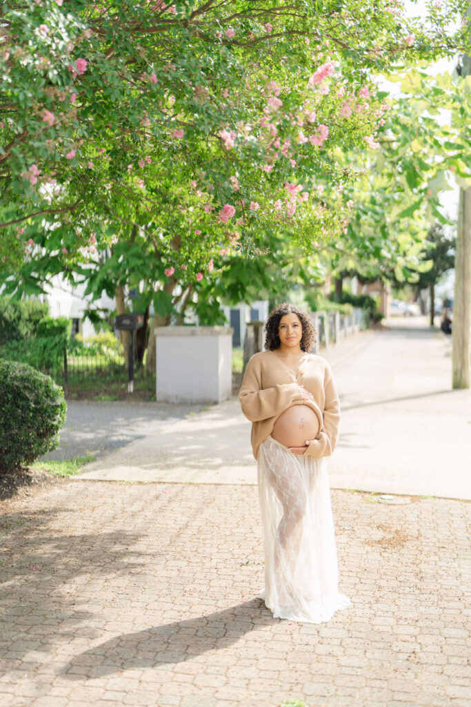 A pregnant woman holds her belly and smiles standing under a large green tree with pink flowers.